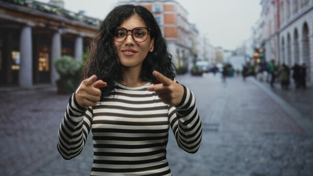 Woman points finger to camera on a cobblestone street, wearing glasses and striped shirt, smiling with curly brunette hair; confidence playfulness.