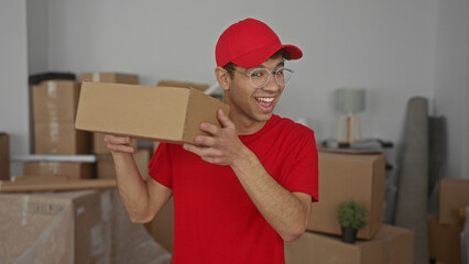 Man holding cardboard box and smiling in building wearing red cap and glasses amid stacked moving boxes; joy.