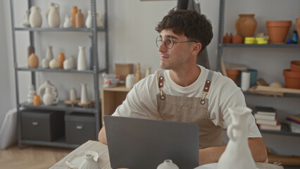 Man with hands clasped and laptop at pottery studio with shelves of ceramics and clay pots, wearing apron and round glasses; quiet contemplation.