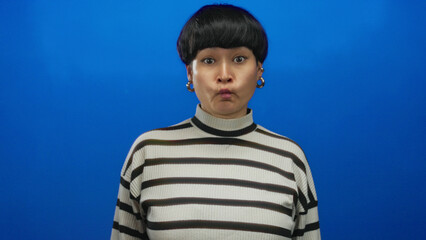 Woman making funny face on blue background wearing striped sweater and earrings, expressing...