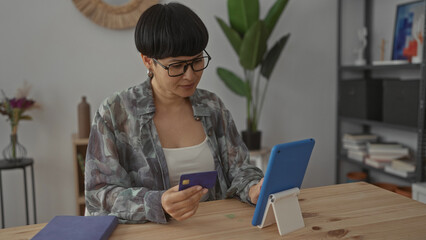 Woman using tablet for online shopping in modern living room holding credit card with focused expression surrounded by cozy home decor and natural light.