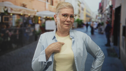 Woman finger pointing to chest on busy street terrace, short cropped haircut, eyeglasses, light shirt, hand on hip, direct gaze; confidence.