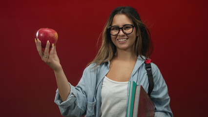 Hispanic teen woman wearing glasses and backpack holding apple and textbooks in red studio; youthful confidence.