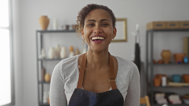 Woman artisan wearing denim apron smiling with visible teeth in studio with pottery shelves and clay tools on racks; creative joy.
