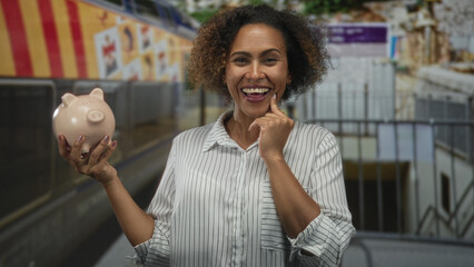 Woman holding piggy bank, finger to cheek, near train in station building, smiling; financial...