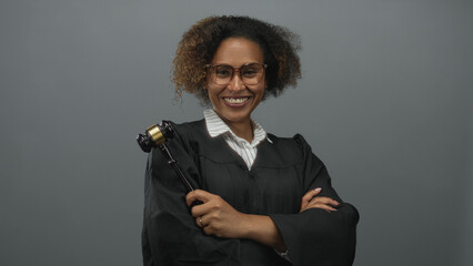 Woman judge wearing robe and glasses smiling while holding gavel and arms crossed in grey studio;...