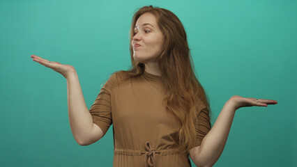 Woman with long red hair and freckles displaying open palms and a puzzled expression in a teal studio; uncertainty.