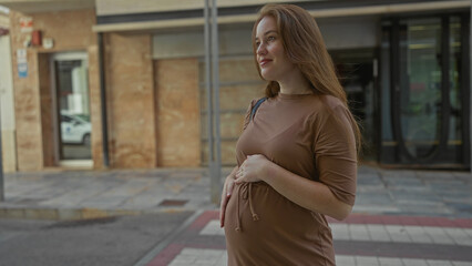 Fototapeta premium Young caucasian pregnant woman gently holding belly on city street with blurred storefronts and tiled pavement under soft daylight; serenity.