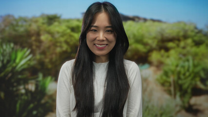 Young chinese woman smiling outdoors in a lush green park, surrounded by vibrant nature and clear blue sky, conveying a sense of tranquility and happiness.