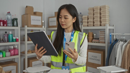 Young woman in reflective uniform using tablet and phone in charity center with donation boxes and food supplies on shelves.