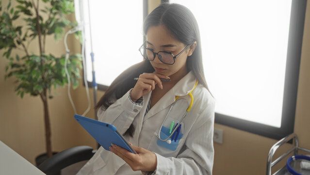 Young chinese woman doctor with stethoscope and glasses using digital tablet in hospital room, analyzing medical data indoors in professional healthcare setting.