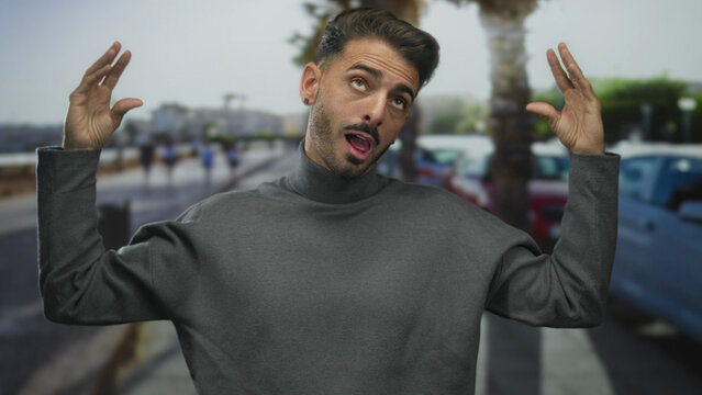 Young hispanic man wearing a grey turtleneck gestures with raised hands on street lined with palm trees; confusion.