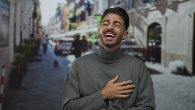 Man holds hand on chest and laughs broadly on narrow sunlit city street lined with blurred buildings; joy.