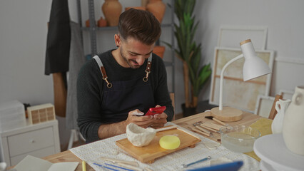 Man in apron holds red smartphone tapping screen beside clay bowl on wooden table with pottery tools in workshop; focused creativity.