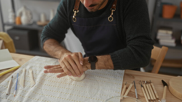 Man kneads clay with bare hands over a wooden worktable surrounded by pottery tools in studio; creativity. - Powered by Adobe