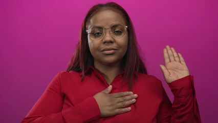 African american woman in studio against pink wall places hand on chest and raises other hand;...