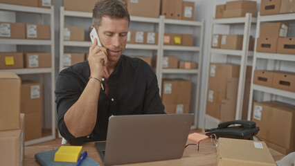 Man holding phone to ear and typing on laptop in a storage building office surrounded by parcels...
