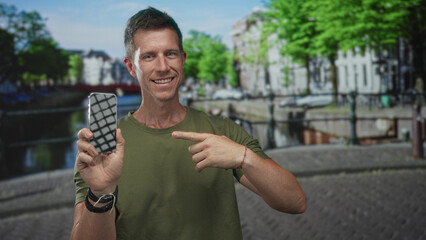 Man points finger at smartphone on street in amsterdam canal quay while smiling and holding phone; confidence connectivity travel.