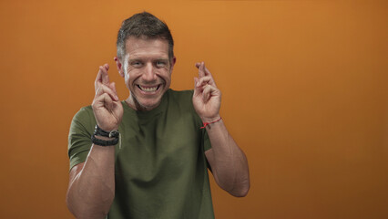 Man with fingers crossed, smiling and showing hands and forearms in orange studio wearing green t shirt and bracelets; hopeful anticipation luck.