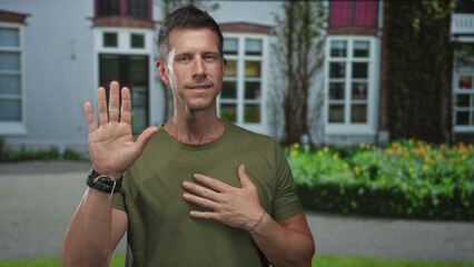 Young man with raised left hand palm out and right hand on chest at residential building entrance outdoors; sincerity.