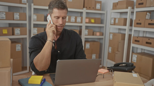 Man holding phone to ear and typing on laptop in a storage building office surrounded by parcels and shelving; focused productivity. - Powered by Adobe