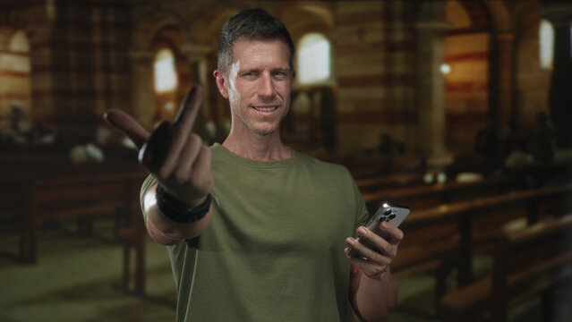 Man shows middle finger while holding smartphone inside church building, wearing green t shirt and wrist bracelets, direct stare; defiance provocation.