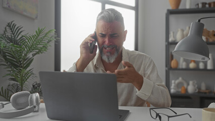 Man talking on phone with hand raised at desk near window while using laptop in a building office; work stress.