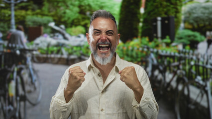 Bearded man clenches fists and shouts with bare hands raised on a park street lined with bicycles; triumph.