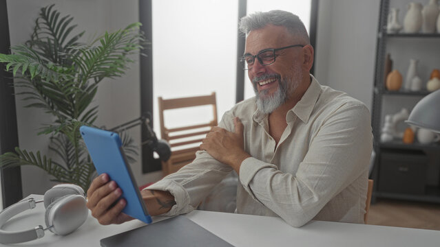 Man smiling and waving hand while holding blue tablet in a building home office with plant and headphones on desk; connection happiness.