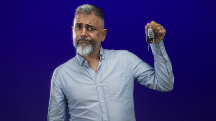 Man holding car keys up with visible hand in studio against deep blue background while raising eyebrow and smirking; skepticism.