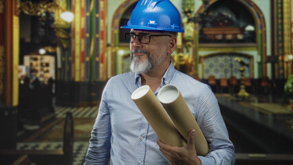 Man architect holding rolled plans and wearing blue hardhat in building church interior; heritage...
