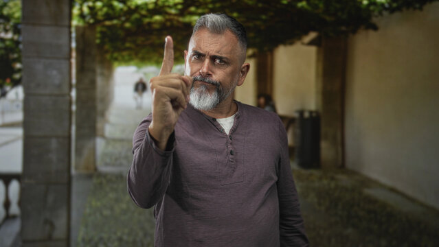 Man with gray beard pointing index finger toward camera in building entrance by house doorway outdoors; stern warning.
