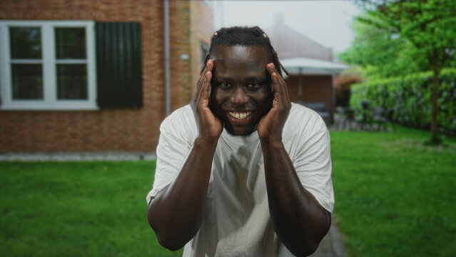 Young man covering face with hands at house entrance, smiling toward camera while leaning forward in a casual pose; playful joy. - Powered by Adobe