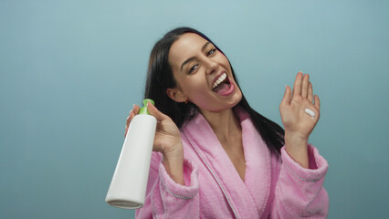 Hispanic woman in a pink bathrobe enthusiastically holds skincare lotion against a blue background suggesting beauty routine.