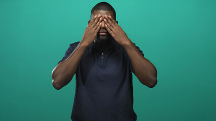 Man covers eyes then opens palms toward camera in studio against teal backdrop, then smiles in reveal gesture; playful joy.