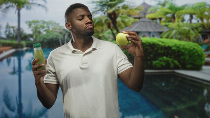 Man holding a green apple and juice bottle in bare hands beside a pool and resort building while...