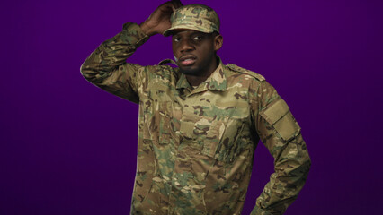 Soldier in camouflage uniform touching cap with right hand, adjusting brim while looking alert in studio; duty focus.