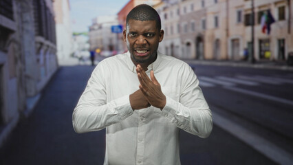 Obraz premium Man rubbing hands and grimacing in a white shirt on a city street with blurred buildings and a crosswalk visible; frustration tension.