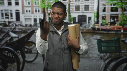Man holding rolled blueprints and raising middle finger, hand visible, on a canal street with...