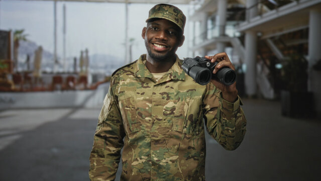 Soldier man holding binoculars near shoulder, smiling in an airport terminal; pride duty vigilance service.