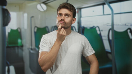 Young hispanic man touching temple with finger inside bus with green seats on street; thoughtful reflection curiosity.