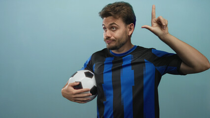 Young hispanic man holding a soccer ball, hand forming l sign to head, wearing blue and black...