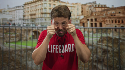 Man lifeguard in red shirt rubbing eyes with fists and whistle visible at building ruins; tired...