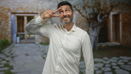 Man making v sign with fingers next to eye at building entrance outside a stone house, smiling and wearing white shirt while looking upward; playful greeting.