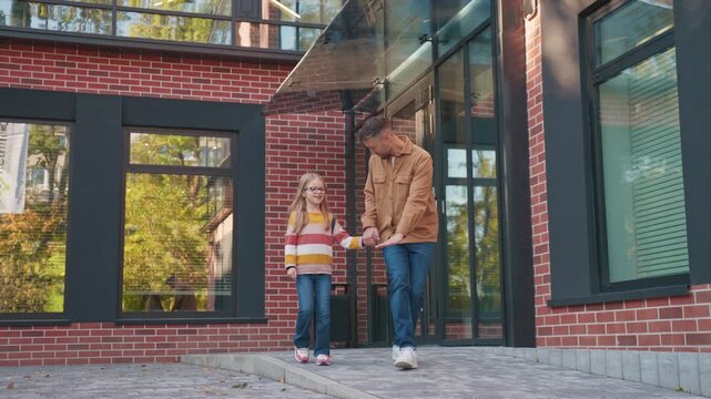 Caucasian father holding hand of daughter while pointing ahead near school entrance. Adult male showing direction to girl beside brick walls. Parent guiding child along walkway before morning drop off