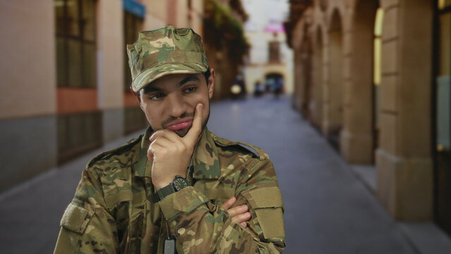 Young hispanic man in green camouflage military uniform with hand on chin contemplating on old town street; inner doubt.