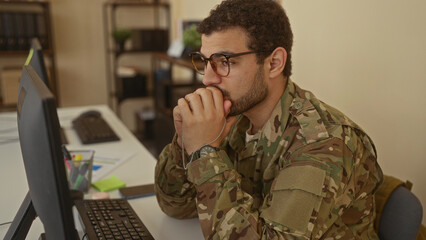 Man holding military dog tags, hand on chin in pensive gesture at a computer desk in a building; reflection.