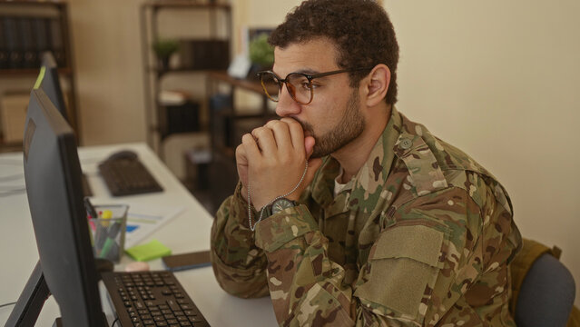 Man holding military dog tags, hand on chin in pensive gesture at a computer desk in a building; reflection.