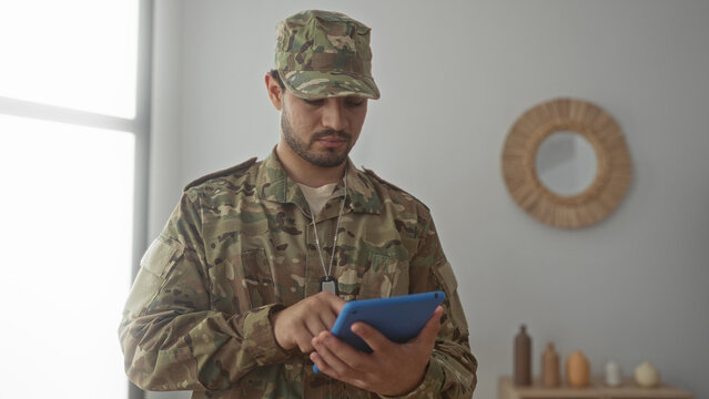 Hispanic man taps tablet in living room while wearing camouflage uniform with dog tags displayed; concentration.
