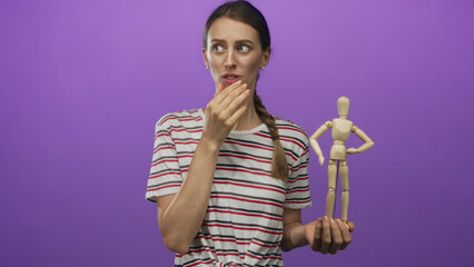 Woman holds wooden mannequin and touches lips in studio with purple backdrop wearing striped shirt and braided ponytail; doubt pondering.